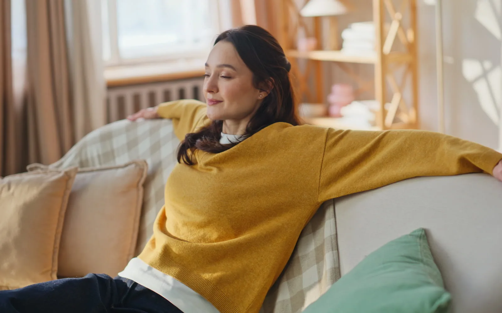 A woman sitting back on a sofa in a sunlit living room, smiling to herself, relaxed and unhurried