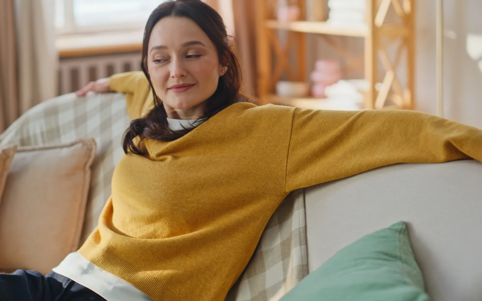 A woman relaxing back on a sofa in a sunlit room, smiling warmly