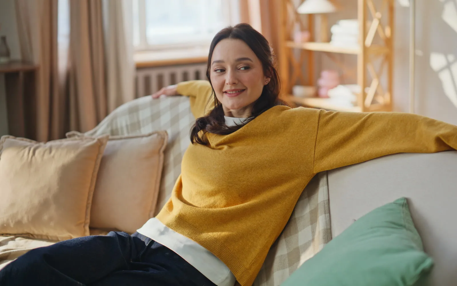A woman relaxing on a sofa at home, smiling to herself in the afternoon light